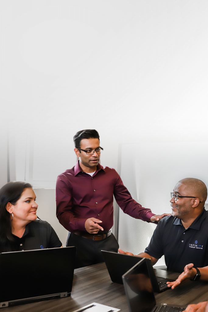 Infinity Tech Consulting professional in burgundy shirt discussing project with two colleagues at conference table with laptops